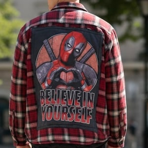 May include: A red, black, and white plaid long-sleeve button-down shirt featuring a large rectangular patch on the back. The patch displays a graphic of the character Deadpool making a heart shape with hands, with the text BELIEVE IN YOURSELF.