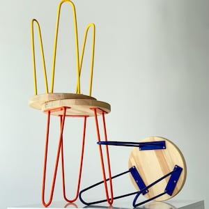 Può includere: Due sgabelli moderni con sedute in legno e gambe in metallo colorato. Uno sgabello ha gambe curve gialle e supporti arancioni, mentre l'altro ha gambe blu. Le sedute sono rotonde e in legno chiaro.