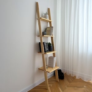 以下が含まれることがあります： 4段の棚が付いた、明るい色の木製の立てかけ式ラダーシェルフ。 棚には本、小さな鉢植え、装飾品が置かれています。 ラダーは白い壁に立てかけられており、白いカーテンのある窓の近くに配置されています。
