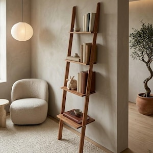 May include: A tall, leaning wooden ladder shelf with four horizontal shelves. The shelves hold various books, a small ceramic bowl, and a white rabbit figurine. The structure is made of medium-toned wood and rests against a light-coloured wall.