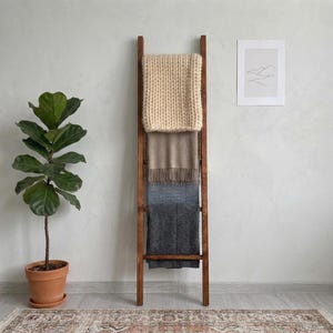 May include: A tall, dark brown wooden ladder leaning against a wall, holding four folded blankets in shades of cream, beige, light grey, and dark charcoal. The cream blanket features a chunky knit texture, while the others have smooth finishes.