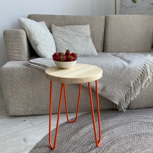 May include: A small wooden table with a round top and orange metal legs. The table is sitting on a gray rug in front of a gray couch with pillows and a throw blanket.