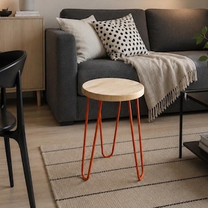 May include: A small round stool with a light-colored natural wood top and three thin, bright orange metal hairpin legs. The stool stands on a light-colored floor, featuring a simple design with a smooth, circular wooden seat and metal legs.