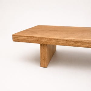 Könnte beinhalten: Ein rechteckiger Holzständer mit einer hellbraunen Oberfläche aus natürlicher Holzmaserung. Die Plattform wird von einem einzelnen vertikalen Bein gestützt, das mittig darunter angebracht ist und eine erhöhte Fläche mit glatter Oberseite bildet.
