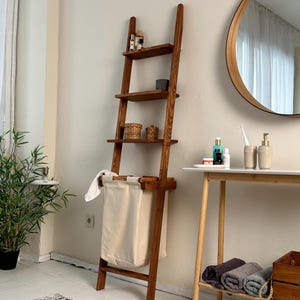 May include: A tall, leaning wooden ladder shelf with a rich brown finish, featuring four horizontal shelves for storage and a large, cream-coloured fabric laundry bag integrated into the bottom section. The structure is made of wood.