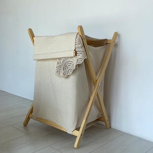 May include: A natural-toned laundry hamper with a wooden frame and a beige fabric liner. A matching fabric cover with a lace trim is draped over the top. The hamper is set against a white wall and light-colored floor.