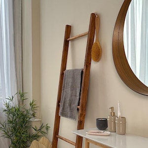 May include: A tall, dark brown wooden ladder leaning against a wall, featuring a grey towel draped over a rung and a round wooden-handled bath brush hanging from the top. The ladder has four horizontal rungs and a smooth, polished wood finish.
