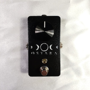 Può includere: Pedale per effetti per chitarra nero con una manopola bianca e una luce LED bianca. Il pedale ha una grafica bianca di tre lune e il testo "OSTARA" sul davanti.