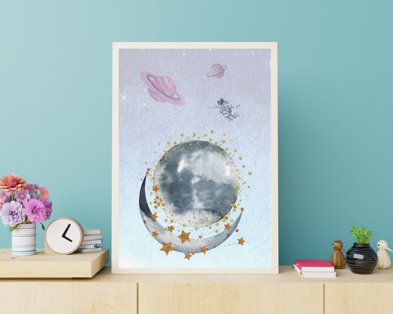 This is a picture of watercolor full and cresnet moon, two Saturns, astronaut and gold stars and on a light purple textured background. The picture is in a wooden photo frame and is propped up against a white wall over a wooden table.