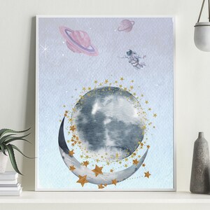 This is a picture of watercolor full and cresnet moon, two Saturns, astronaut and gold stars and on a light purple textured background. The picture is in a wooden photo frame and is propped up against a white wall over a wooden table.