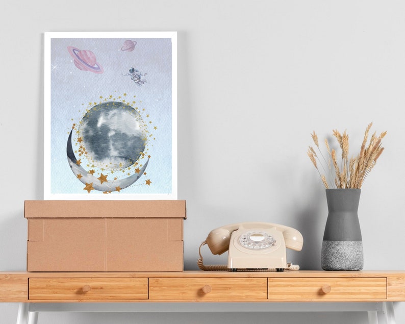 This is a picture of watercolor full and cresnet moon, two Saturns, astronaut and gold stars and on a light purple textured background. The picture is in a wooden photo frame and is propped up against a white wall over a wooden table.