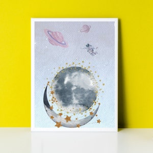 This is a picture of watercolor full and cresnet moon, two Saturns, astronaut and gold stars and on a light purple textured background. The picture is in a wooden photo frame and is propped up against a white wall over a wooden table.