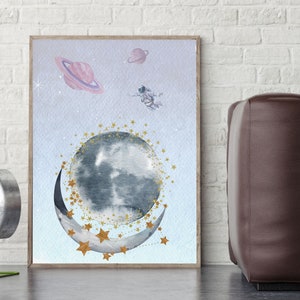 This is a picture of watercolor full and cresnet moon, two Saturns, astronaut and gold stars and on a light purple textured background. The picture is in a wooden photo frame and is propped up against a white wall over a wooden table.