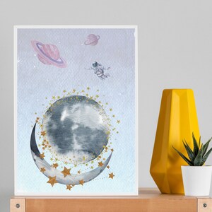 This is a picture of watercolor full and cresnet moon, two Saturns, astronaut and gold stars and on a light purple textured background. The picture is in a wooden photo frame and is propped up against a white wall over a wooden table.
