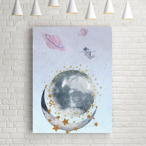 This is a picture of watercolor full and cresnet moon, two Saturns, astronaut and gold stars and on a light purple textured background. The picture is in a wooden photo frame and is propped up against a white wall over a wooden table.