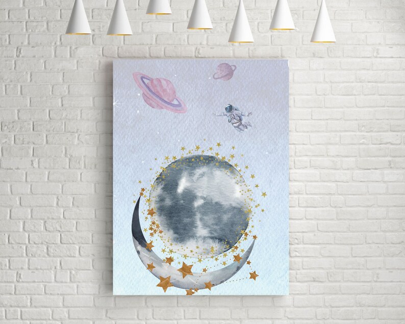 This is a picture of watercolor full and cresnet moon, two Saturns, astronaut and gold stars and on a light purple textured background. The picture is in a wooden photo frame and is propped up against a white wall over a wooden table.