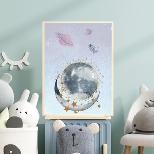 This is a picture of watercolor full and cresnet moon, two Saturns, astronaut and gold stars and on a light purple textured background. The picture is in a wooden photo frame and is propped up against a white wall over a wooden table.