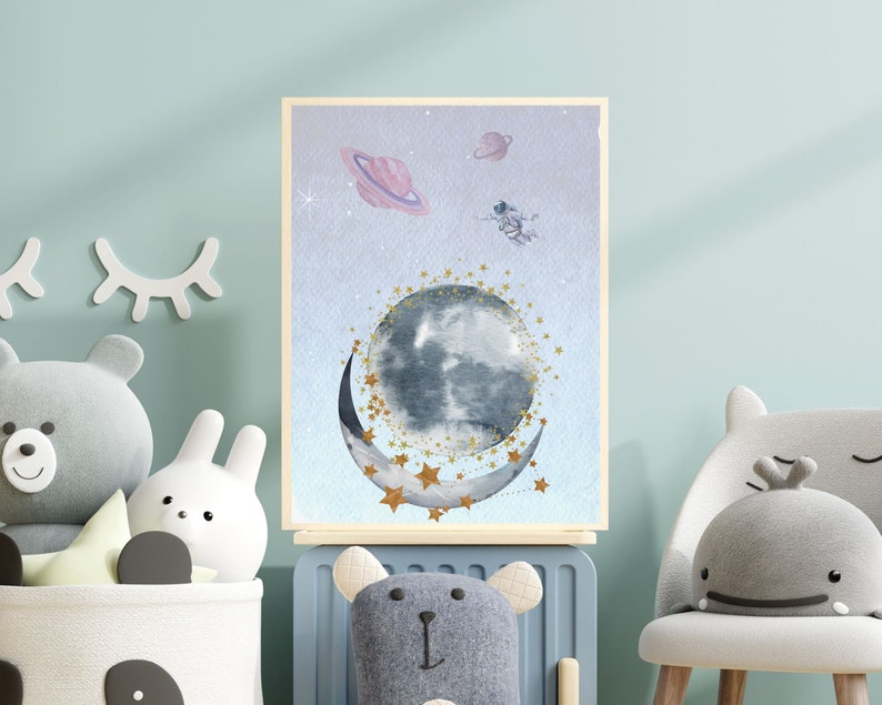 This is a picture of watercolor full and cresnet moon, two Saturns, astronaut and gold stars and on a light purple textured background. The picture is in a wooden photo frame and is propped up against a white wall over a wooden table.