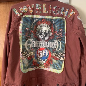 May include: A brown denim jacket with a large Grateful Dead patch on the back. The patch features a portrait of Jerry Garcia, the band's logo, and the text "Celebrating 50 Years 1965-2015". The words "Lovelight" are sewn on the top of the patch in colourful fabric.