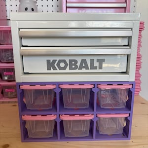 May include: A white Kobalt metal tool chest with two drawers featuring silver handles, stacked on top of a purple plastic storage organizer containing six clear bins with pink lids arranged in two rows of three.