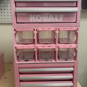 May include: A pink metal storage cabinet featuring two top drawers with silver handles, a middle section with six clear plastic bins, and three bottom drawers with silver handles. The brand name KOBALT is printed in white on the top and bottom drawers.