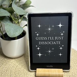 Puede incluir: Un lector electrónico Kindle negro con una funda blanca y una pantalla que muestra el texto "GUESS I'LL JUST DISSOCIATE" con estrellas blancas sobre un fondo negro. El Kindle está en un soporte de madera.