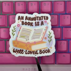 May include: A die-cut sticker featuring an open book resting on a closed book with floral patterns. The text reads AN ANNOTATED BOOK IS A WELL-LOVED BOOK. The design includes green leafy sprigs, small pink flowers, and yellow star accents on a light purple background.