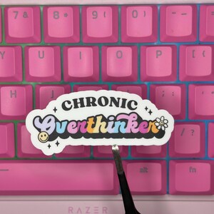 May include: A die-cut vinyl sticker featuring the text "CHRONIC Overthinker" in a bold, retro-inspired font. The word "CHRONIC" is black, while "Overthinker" features a multi-colored gradient of purple, pink, blue, and orange. Includes a small smiley face and a flower icon.