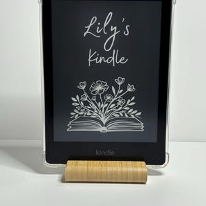 May include: A black Kindle e-reader with a clear case and a personalised inscription that reads "Lily's Kindle". The inscription is in a white, cursive font. The e-reader is sitting on a wooden stand.