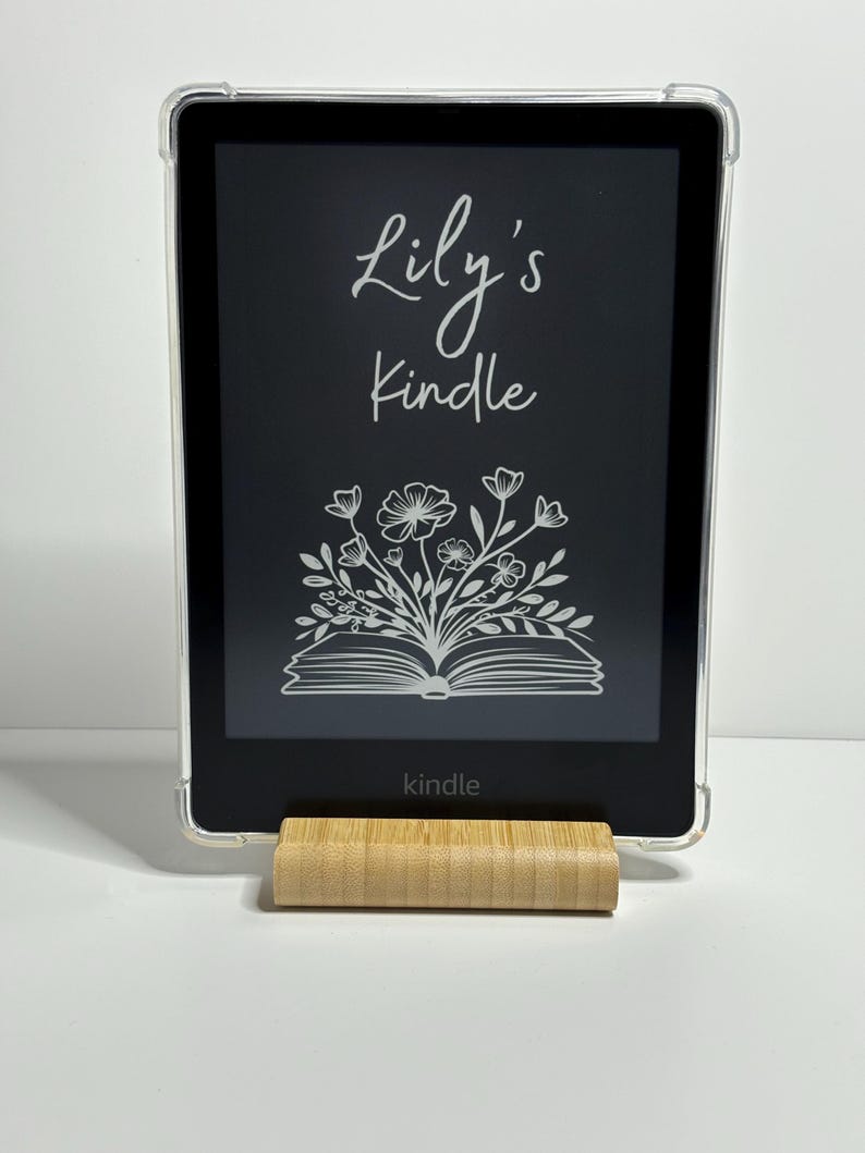 May include: A black Kindle e-reader with a clear case and a personalized inscription that reads "Lily's Kindle". The inscription is in a white, cursive font. The e-reader is sitting on a wooden stand.