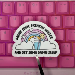 May include: A die-cut sticker featuring a pastel rainbow, white clouds, and a glass of blue water with a straw and a smiley face. The text reads DRINK SOME FREAKIN WATER AND GET SOME DAMN SLEEP in black capital letters against a white background.