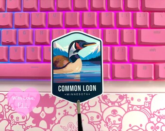 May include: A sticker featuring a Common Loon in a lake scene with mountains in the background. The sticker has the text "COMMON LOON - MINNESOTA." The sticker is held in front of a pink keyboard.