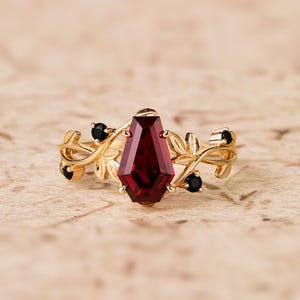 May include: A gold ring with a large, dark red gemstone set in a coffin-shaped setting. The ring is adorned with delicate gold leaves and small black gemstones.
