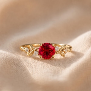 Op de afbeelding: Een gouden ring met een ronde, felrode centrale steen. De band is aan beide zijden ontworpen met delicate, gebeeldhouwde bladmotieven, geaccentueerd door kleine, heldere, rond geslepen stenen die in het metalen loof rond de edelsteen zijn gezet.