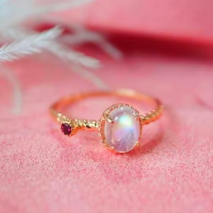 Op de afbeelding: Een gouden ring met een witte ovale maansteen en een klein rood edelsteentje. De ring heeft een gedraaide band.