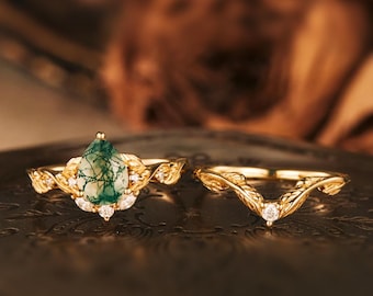 May include: Two gold rings. The engagement ring features a pear-shaped green moss agate stone, surrounded by small white diamonds. The band has a leaf design. The wedding band is a curved gold band with a single white diamond.