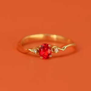 May include: A gold ring with a red oval gemstone and two smaller white gemstones. The ring has a delicate, twisted band.