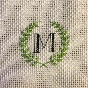 May include: A white fabric square with a green wreath embroidered around a black and white letter M.