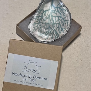 May include: A decorative oyster shell dish with a silver-colored rim and a teal branch design inside, presented in a small brown box. The box has a label that reads "Nautical By Desiree Est. 2021 Made by hand, inspired by the ocean."