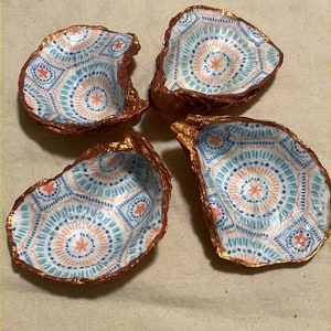 Pode incluir: Quatro conchas de ostra decorativas. Cada concha tem um interior pintado com um padrão geométrico azul, laranja e branco. As bordas são adornadas com uma cor dourada metálica, criando uma estética costeira e boêmia.