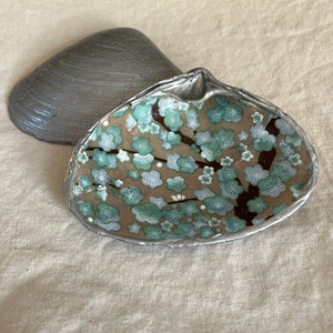 May include: Two decorative clam shells. One shell is painted a metallic gray. The other shell is a dish with a floral design of teal blossoms on brown branches against a tan background. The rim of the dish is silver.