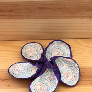 Pode incluir: Seis conchas de ostra decorativas com exterior roxo e interior estampado. O design interior apresenta um padrão repetitivo de linhas e estrelas azuis, pêssego e brancas. As conchas estão dispostas sobre uma superfície de madeira.