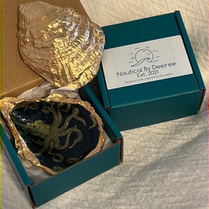 Pode incluir: Uma caixa turquesa aberta com um prato decorativo de concha de ostra com um desenho de polvo dourado. Uma segunda concha de ostra pintada de ouro está ao lado da caixa. A caixa tem um rótulo que diz "Nautical By Desiree Est. 2021 Made by hand, inspired by the ocean."