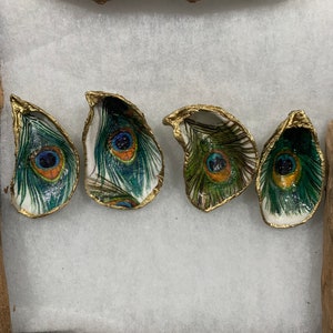 May include: Four oyster shells with peacock feather designs. The shells have gold-painted edges and feature vibrant blue, green, and orange details. The shells are arranged on a white surface, with driftwood accents.