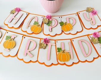May include: A pink and orange Happy Birthday banner with pumpkin and flower accents. The banner is made of felt and has a scalloped edge.