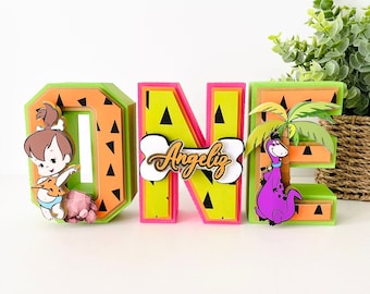 Puede incluir: Decoraciones para el primer cumpleaños con los personajes de la caricatura "Los Picapiedra". Las letras son verdes, amarillas y naranjas con acentos negros y blancos. Las letras están decoradas con una caricatura de Pebbles Picapiedra, un hueso con el nombre "Angeliz" escrito en él y un dinosaurio morado.