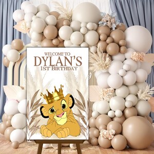 May include: A welcome sign for a first birthday party with a brown and beige balloon backdrop. The sign features a cartoon lion cub wearing a gold crown and the text "Welcome to Dylan's 1st Birthday".