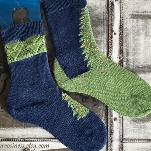 Pode incluir: Um par de meias de Natal de malha. Uma meia é azul marinho com um calcanhar verde e um desenho de árvore de Natal verde. A outra meia é verde com um punho azul marinho e um desenho geométrico verde. As meias são exibidas em uma superfície de madeira.