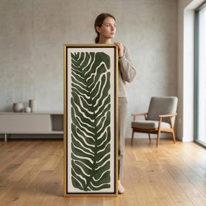 May include: A framed abstract art piece featuring a dark green leaf design on a cream background. The artwork is encased in a gold frame and is displayed vertically. The overall aesthetic is modern and minimalist.