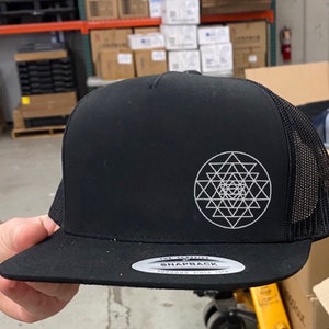 May include: Black trucker hat with a white geometric design on the front. The hat has a mesh back and a flat brim. The text "THE CLASSICS SNAPBACK" is on the brim.